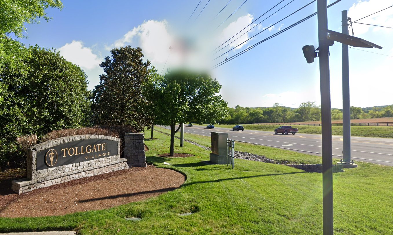 Tollgate Village entrance sign beside a road, with a pole-mounted solar ALPR-style camera at the roadside. Street View imagery dated April 2023.